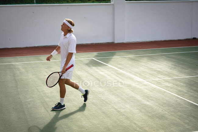 A Caucasian man wearing tennis whites spending time on a court playing tennis on a sunny day, holding a tennis racket — Stock Photo