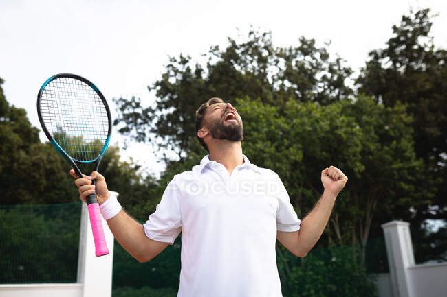 Змішана гонка чоловік в тенісних білих проводить час на корт, граючи в теніс в сонячний день, тримаючи тенісну ракетку і святкуючи — стокове фото