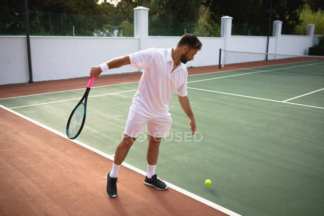 Ein Mann mit gemischter Rasse in weißen Tennisanzügen verbringt an einem sonnigen Tag Zeit auf einem Tennisplatz und bereitet sich darauf vor, einen Ball zu schlagen — Stockfoto