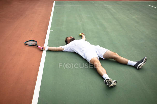 Um homem de raça mista vestindo brancos de tênis passando tempo em uma quadra jogando tênis em um dia ensolarado, deitado em um chão, segurando uma raquete de tênis e uma bola — Fotografia de Stock