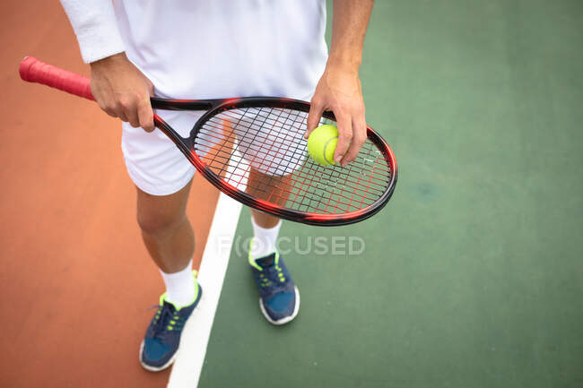 Vista a meio da seção do homem usando brancos de tênis passando tempo em um campo de ténis em um dia ensolarado, segurando uma raquete de tênis e uma bola — Fotografia de Stock