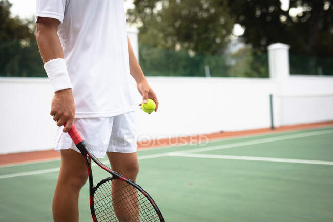 Средняя часть мужчины в теннисных белках проводит время на корте, играя в теннис в солнечный день, держа теннисную ракетку, готовясь ударить по мячу — стоковое фото