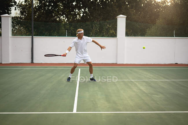 Un homme caucasien portant des blancs de tennis passe du temps sur un court de tennis par une journée ensoleillée, tenant une raquette de tennis, se préparant à frapper une balle — Photo de stock