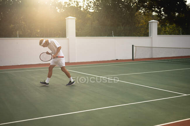Ein kaukasischer Mann in weißen Tennisanzügen, der an einem sonnigen Tag mit einem Tennisschläger auf einem Tennisplatz spielt — Stockfoto