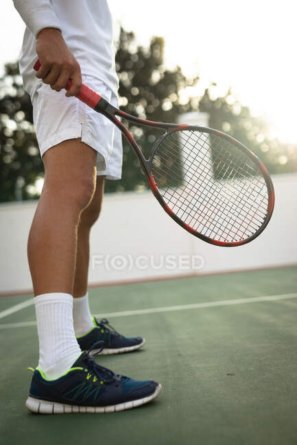 Sección media del hombre que usa ropa blanca de tenis que pasa tiempo en una cancha jugando al tenis en un día soleado, sosteniendo una raqueta de tenis - foto de stock