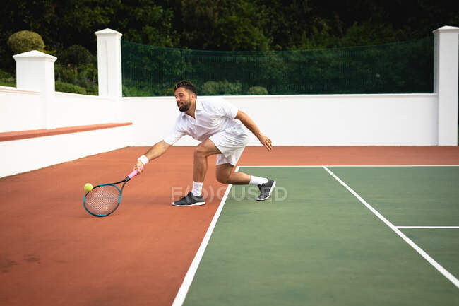 Ein Mann mit gemischter Rasse in weißen Tennisanzügen verbringt an einem sonnigen Tag seine Zeit auf einem Tennisplatz und schlägt mit einem Tennisschläger auf einen Ball — Stockfoto