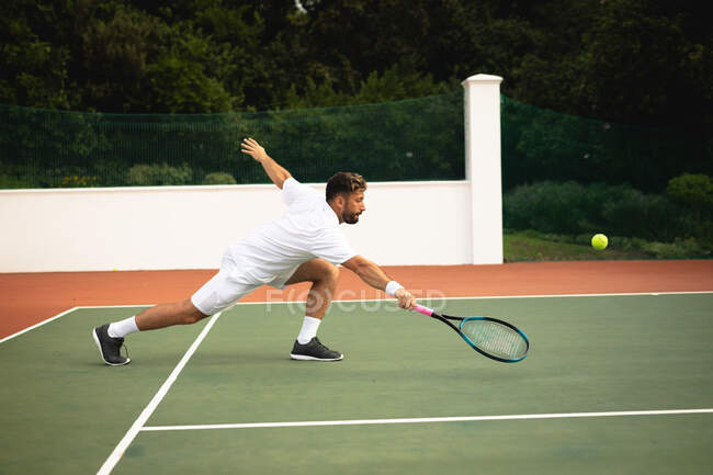 Un homme de race mixte portant des blancs de tennis passe du temps sur un court de tennis par une journée ensoleillée, frappant une balle avec une raquette de tennis — Photo de stock