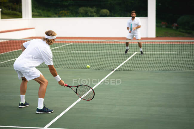 Ein kaukasischer und ein gemischter Rasse Männer in weißen Tennisanzügen verbringen Zeit auf einem Tennisplatz zusammen, spielen Tennis an einem sonnigen Tag, halten Tennisschläger und schlagen einen Ball — Stockfoto