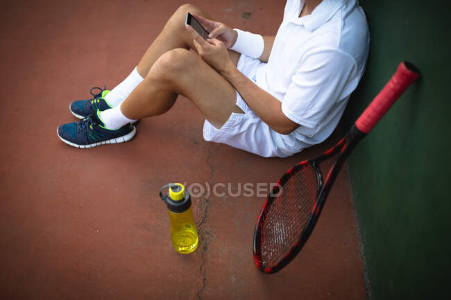 Средний вид на человека в теннисных белках, проводящего время на корте, играющего в теннис в солнечный день, сидящего на земле, используя смартфон, с теннисной ракеткой рядом — стоковое фото
