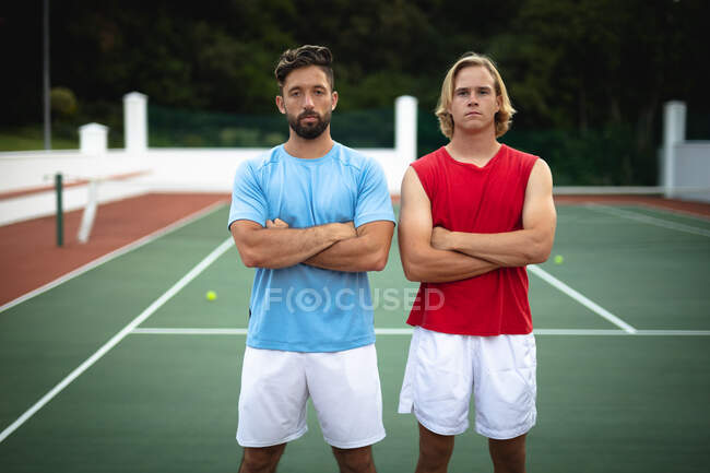 Porträt einer kaukasischen und einer gemischten Rasse, die an einem sonnigen Tag gemeinsam Tennis spielen, die Arme verschränkt in die Kamera starrend — Stockfoto