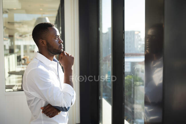 Un uomo d'affari afroamericano, indossa una camicia bianca, lavora in un ufficio moderno, guarda attraverso una finestra, tocca il mento e pensa: — Foto stock