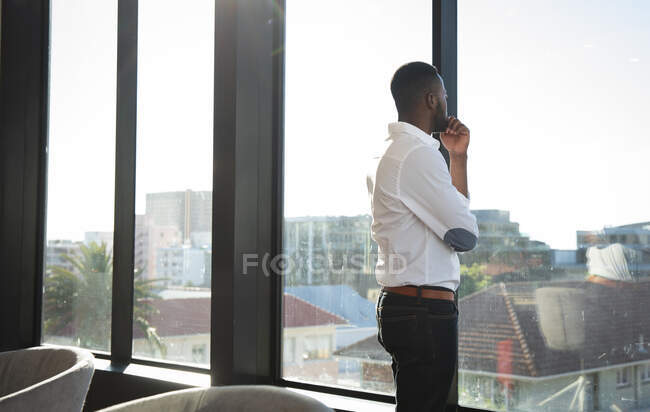 Un hombre de negocios afroamericano, vistiendo una camisa blanca, trabajando en una oficina moderna, mirando por una ventana, tocándose la barbilla y pensando - foto de stock