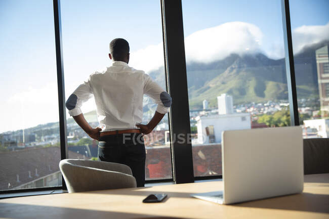 Un hombre de negocios afroamericano, con una camisa blanca, trabajando en una oficina moderna, mirando a través de una ventana, con las manos en la cintura - foto de stock