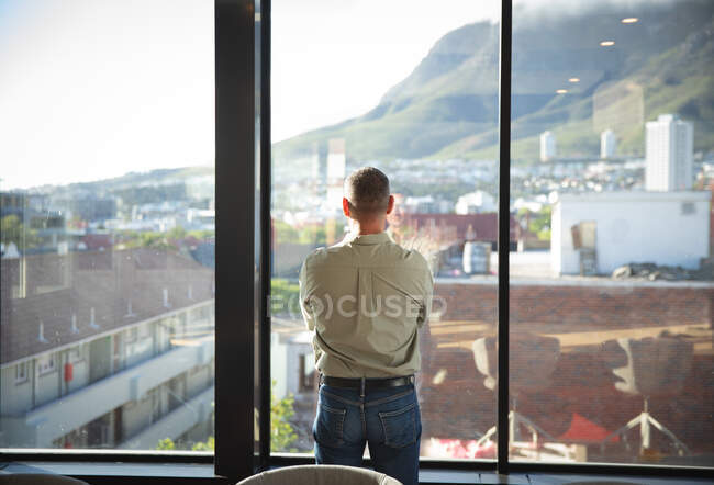 Un homme d'affaires caucasien, portant une chemise grise, travaillant dans un bureau moderne, regardant par une fenêtre, par une journée ensoleillée — Photo de stock