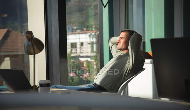Un hombre de negocios caucásico que trabaja en una oficina moderna, sentado en un escritorio, sosteniendo la cabeza, mirando hacia otro lado y pensando, en un día soleado - foto de stock