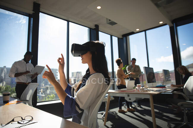 Una mujer de negocios asiática que trabaja en una oficina moderna, con auriculares VR, tocando la pantalla interactiva virtual, con sus colegas trabajando en segundo plano - foto de stock