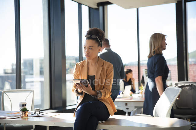 Une femme d'affaires métisse travaillant dans un bureau moderne, assise sur un bureau et utilisant une tablette, avec ses collègues travaillant en arrière-plan — Photo de stock