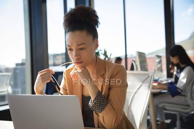 Деловая женщина смешанной расы, работающая в современном офисе, сидящая за столом и используя ноутбук, со своим коллегой, работающим на заднем плане — стоковое фото