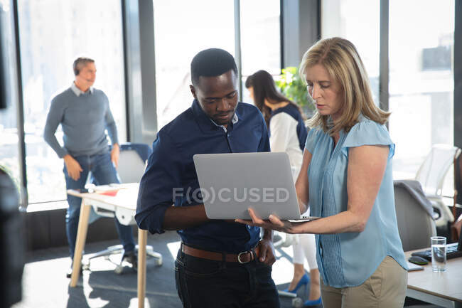 A Caucasian businesswoman and an African American businessman working in a modern office, using a laptop computer and talking, with their colleagues working in the background — Stock Photo