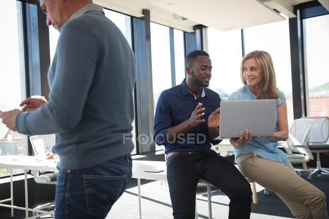 Une femme d'affaires caucasienne et un homme d'affaires afro-américain travaillant dans un bureau moderne, utilisant un ordinateur portable et parlant, avec leur collègue travaillant au premier plan — Photo de stock