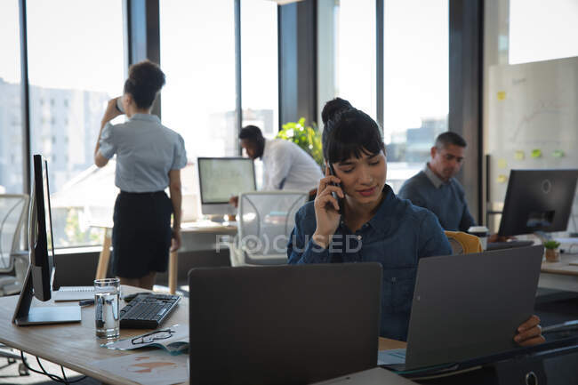 Азійська бізнесменка, яка працює в сучасному офісі, сидить за столом, користується ноутбуком і розмовляє на смартфоні, а її колеги працюють на задньому плані. — стокове фото