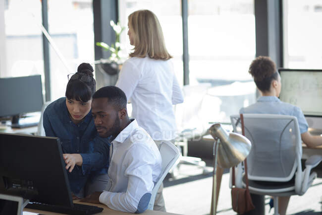 Uma empresária asiática e um empresário afro-americano trabalhando em um escritório moderno, usando um computador e conversando, com seus colegas trabalhando em segundo plano — Fotografia de Stock