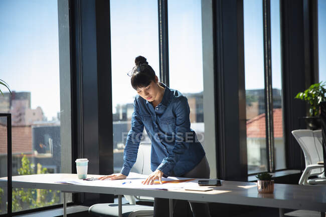 Una mujer de negocios asiática que trabaja en una oficina moderna, de pie junto a un escritorio y mirando planes, en un día soleado - foto de stock