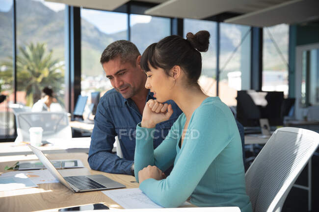 Una empresaria asiática y un hombre de negocios caucásico trabajando en una oficina moderna, usando una computadora portátil y hablando, con sus colegas trabajando en segundo plano - foto de stock