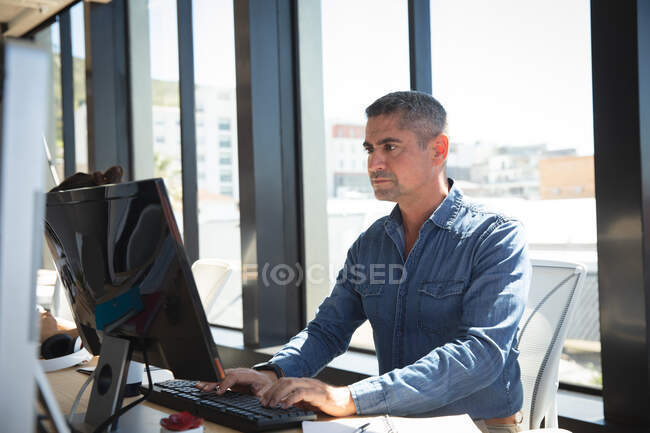 Кавказский бизнесмен, работающий в современном офисе, сидящий за столом и пользующийся настольным компьютером, в солнечный день — стоковое фото