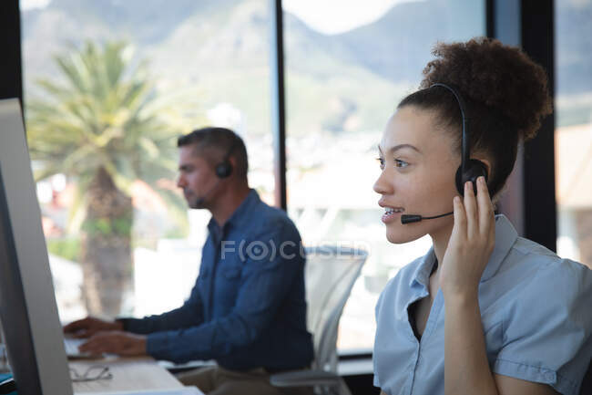 Una mujer de negocios de raza mixta que trabaja en una oficina moderna, sentada en un escritorio, usando una computadora, usando auriculares y hablando, con su colega trabajando en el fondo - foto de stock