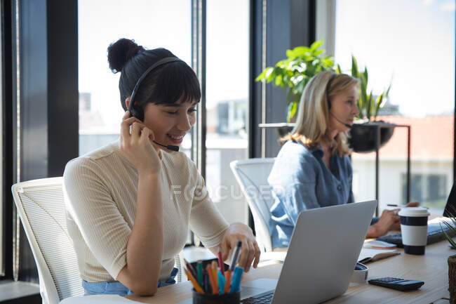 Une femme d'affaires asiatique travaillant dans un bureau moderne, assise à un bureau, utilisant un ordinateur portable, portant un casque et parlant, avec son collègue travaillant en arrière-plan — Photo de stock