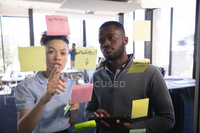 Une femme d'affaires métisse et un homme d'affaires afro-américain travaillant dans un bureau moderne, brainstorming écrit sur un tableau clair avec des notes de service, vu à travers — Photo de stock