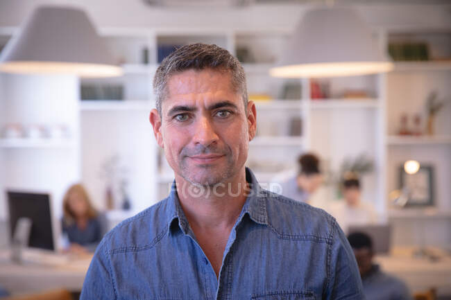Портрет щасливого кавказького бізнесмена, який працює в сучасному офісі, дивиться на камеру і посміхається, з колегами, які працюють на задньому плані. — стокове фото