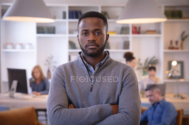 Retrato de un hombre de negocios afroamericano trabajando en una oficina moderna, mirando a la cámara con los brazos cruzados, con sus colegas trabajando en el fondo - foto de stock