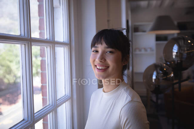 Retrato de una feliz empresaria asiática trabajando en una oficina moderna, de pie junto a una ventana, mirando a la cámara y sonriendo - foto de stock