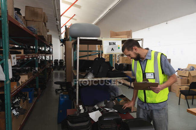 Trabalhador masculino caucasiano num armazém numa fábrica que fabrica cadeiras de rodas, de pé e a inspeccionar peças nas prateleiras, segurando uma prancheta — Fotografia de Stock