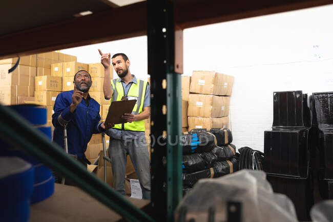 Um trabalhador afro-americano e um supervisor caucasiano em um armazém em uma fábrica de cadeiras de rodas, de pé e falando, segurando uma prancheta — Fotografia de Stock