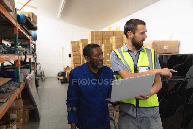 Ein afroamerikanischer Arbeiter und ein kaukasischer männlicher Vorgesetzter in einer Lagerhalle einer Fabrik, die Rollstühle herstellt, stehen und reden, einen Laptop in der Hand — Stockfoto