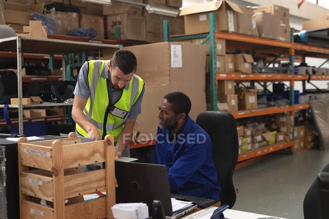 An African American male worker and a Caucasian male supervisor in a storage warehouse at a factory making wheelchairs, standing and talking at a workbench — Stock Photo