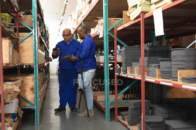 Змішана раса і афроамериканські чоловіки на складі на заводі роблять інвалідні візки, стоять і перевіряють деталі на полицях. — стокове фото