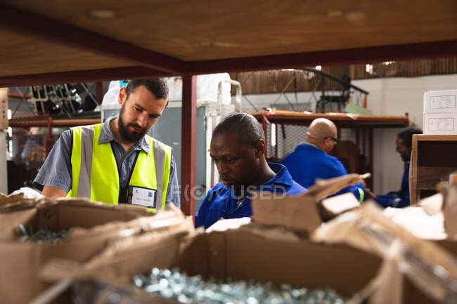 A Caucasian male supervisor and an African American worker in a storage warehouse at a factory making wheelchairs, standing and inspecting parts on shelves — Stock Photo