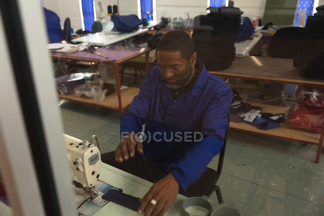 Un trabajador afroamericano en un taller en una fábrica que fabrica sillas de ruedas, sentado en un banco de trabajo, usando una máquina de coser - foto de stock