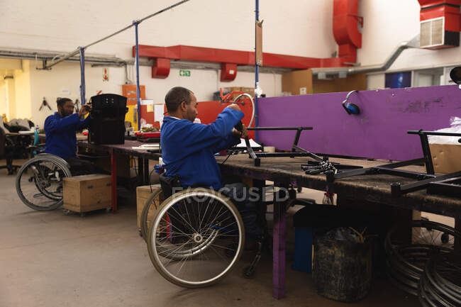 Two disabled African American male workers in a workshop at a factory making wheelchairs, sitting at a workbench assembling parts of a product, sitting in wheelchairs — Stock Photo