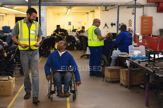 Un trabajador afroamericano discapacitado en una silla de ruedas y un supervisor caucásico hablando en una fábrica haciendo sillas de ruedas, con otros trabajadores trabajando en segundo plano - foto de stock
