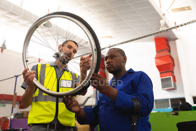 An African American male worker and a Caucasian male supervisor in the workshop at a factory making wheelchairs, standing and inspecting a wheel together, wearing a workwear — Stock Photo