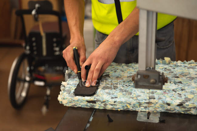 Работник-мужчина в мастерской на фабрике, делающий инвалидные коляски, стоящий на рабочем месте с помощью бензопилы, чтобы разрезать блок пены, отмечая его — стоковое фото