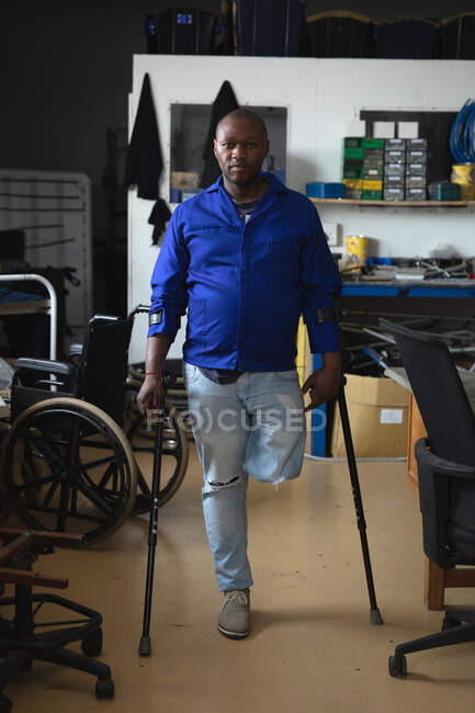 Retrato de un trabajador afroamericano discapacitado con una pierna de pie usando muletas usando ropa de trabajo, en un almacén en una fábrica haciendo sillas de ruedas, mirando a la cámara - foto de stock