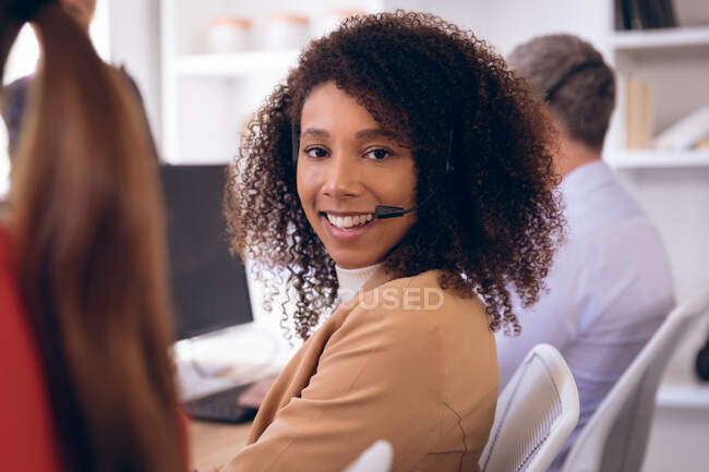 Portrait d'une femme d'affaires métisse travaillant dans un bureau moderne, assise à un bureau, utilisant un ordinateur, portant un casque et parlant, avec son collègue d'affaires travaillant en arrière-plan — Photo de stock