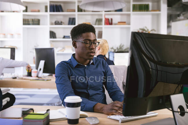 Un uomo d'affari afroamericano che lavora in un ufficio moderno, seduto alla scrivania e usando un computer, con i suoi colleghi d'affari che lavorano sullo sfondo — Foto stock