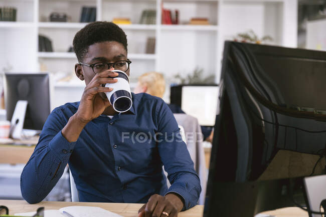 Ein afroamerikanischer Geschäftsmann, der in einem modernen Büro arbeitet, an einem Schreibtisch sitzt und einen Computer benutzt, Kaffee zum Mitnehmen trinkt, während seine Geschäftskollegen im Hintergrund arbeiten — Stockfoto